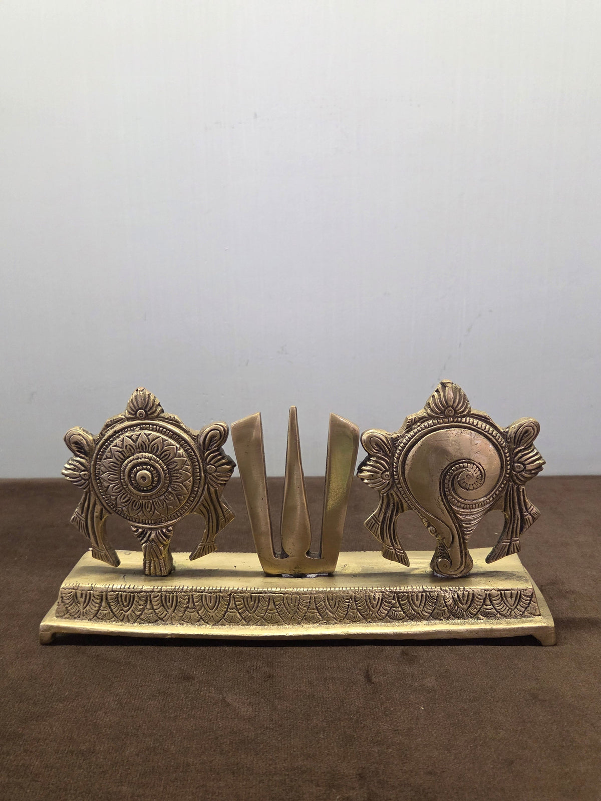 Cast brass sudarshan disc, three-pronged namah and spiral conch standing in a row on an engraved plinth - Peetal Parampara