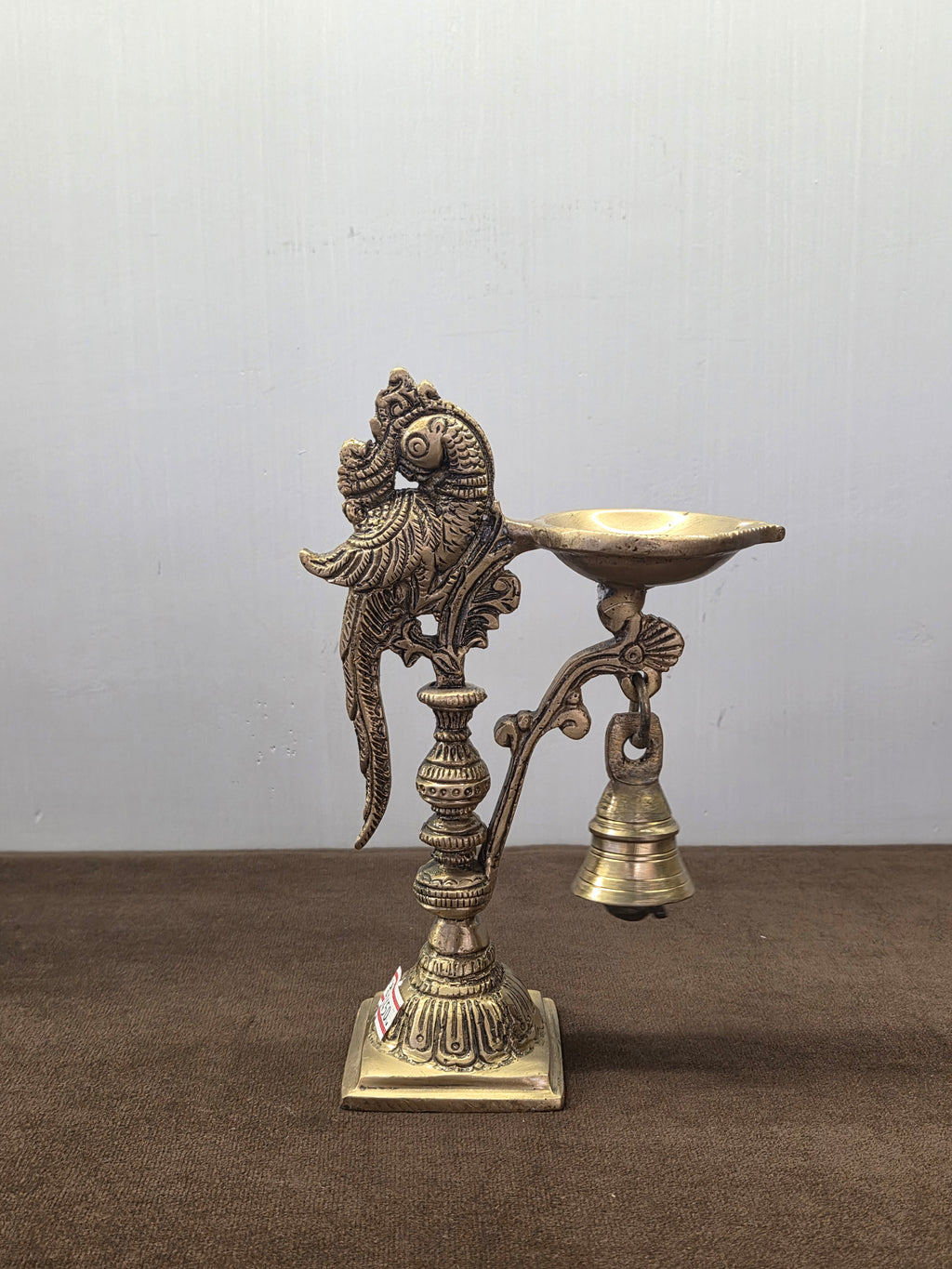 Cast brass parrot diya with an oval oil pan on a side arm, a bell below it and a square stepped base - Peetal Parampara