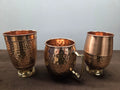 Two hammered copper tumblers on turned brass feet beside a barrel mug with a flat brass strap handle - Peetal Parampara