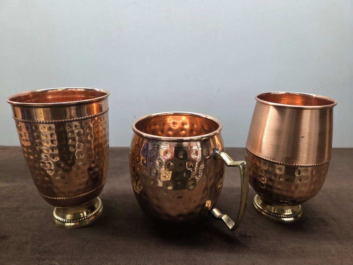Two hammered copper tumblers on turned brass feet beside a barrel mug with a flat brass strap handle - Peetal Parampara