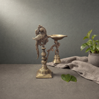 Antique Brass Parrot Diya with Bell on a stone ledge against a grey plaster wall - Peetal Parampara