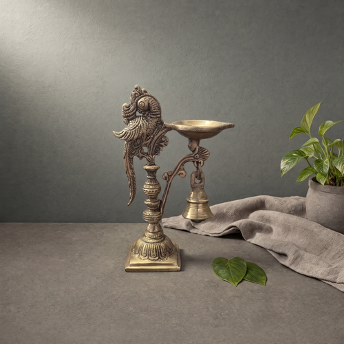 Antique Brass Parrot Diya with Bell on a stone ledge against a grey plaster wall - Peetal Parampara