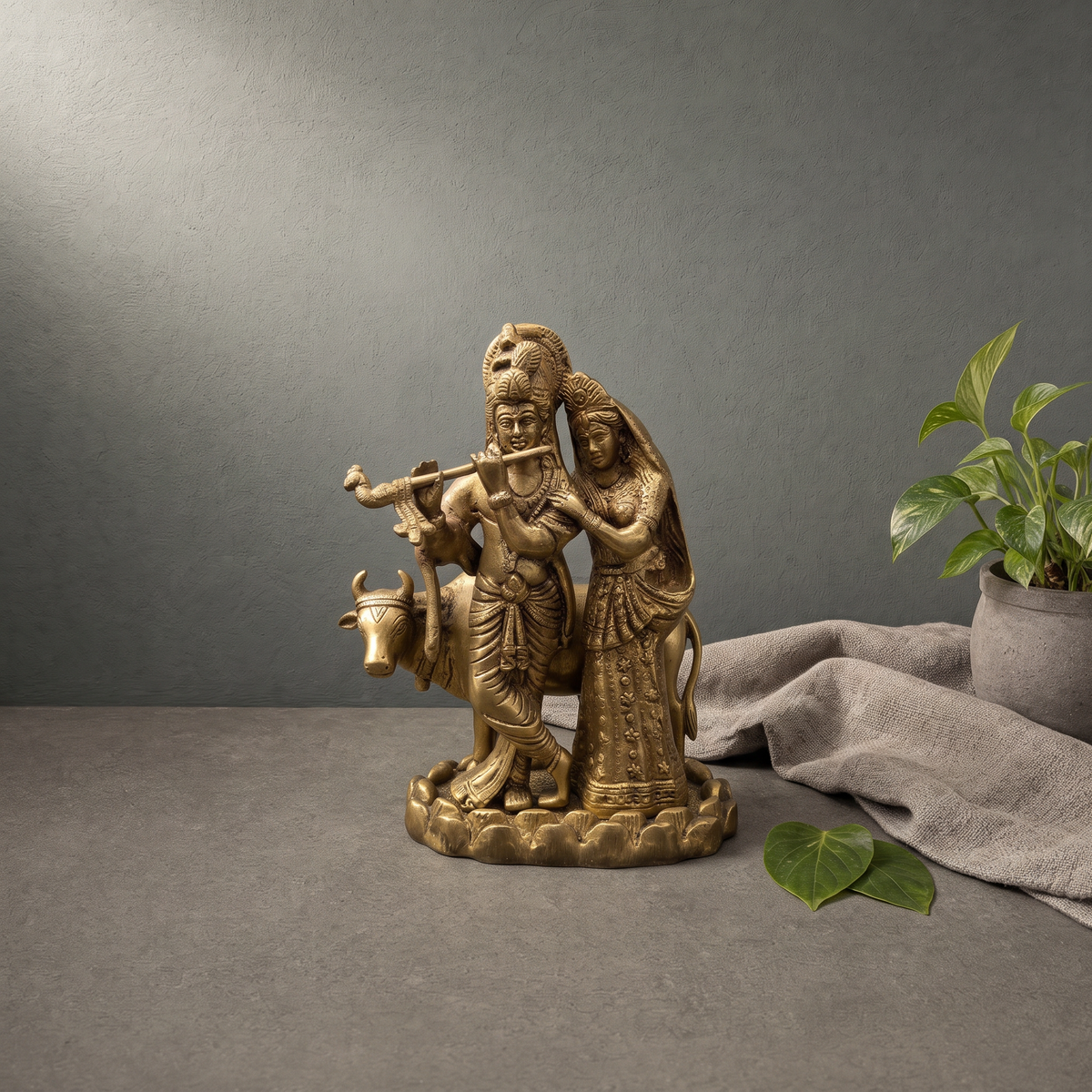 Antique Brass Radha Krishna Murti with Cow on a stone ledge against a grey plaster wall - Peetal Parampara