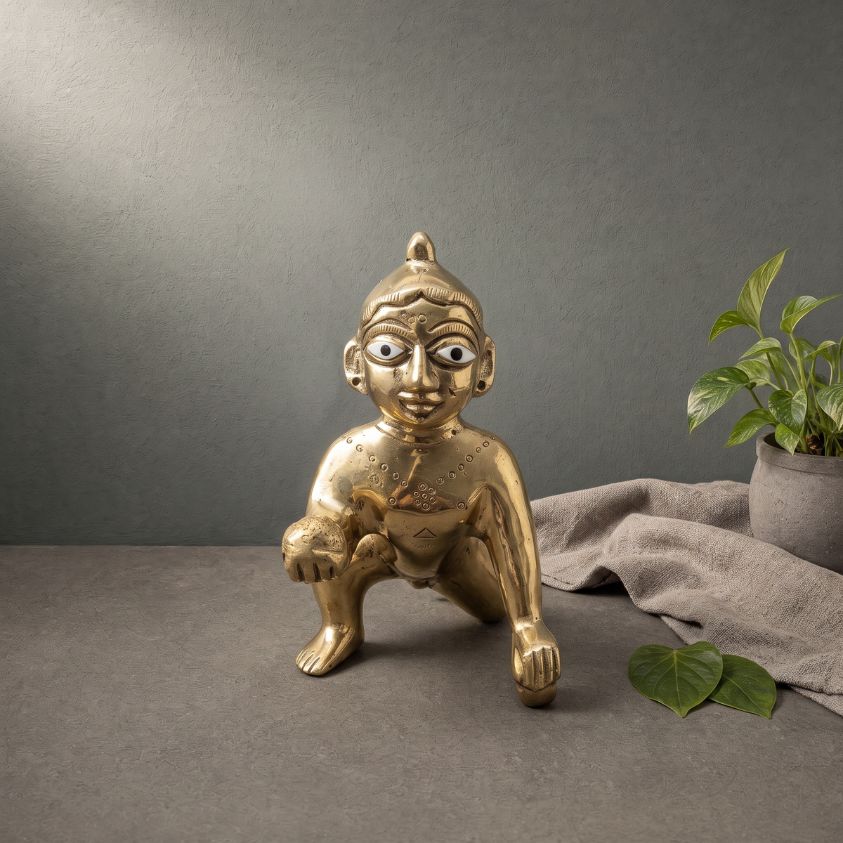 Brass Crawling Laddu Gopal Murti on a stone ledge against a grey plaster wall - Peetal Parampara