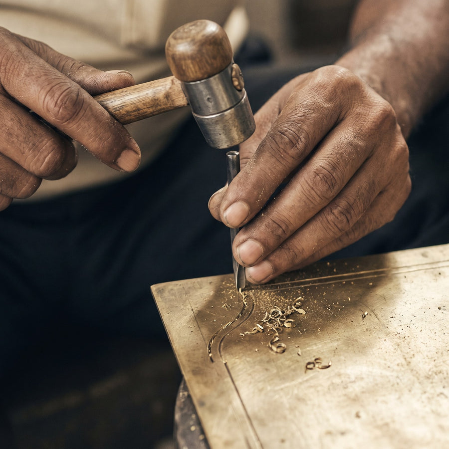 A hand engraving tool and a wooden mallet cutting a line into a flat brass plate, with curls of brass swarf beside the cut.