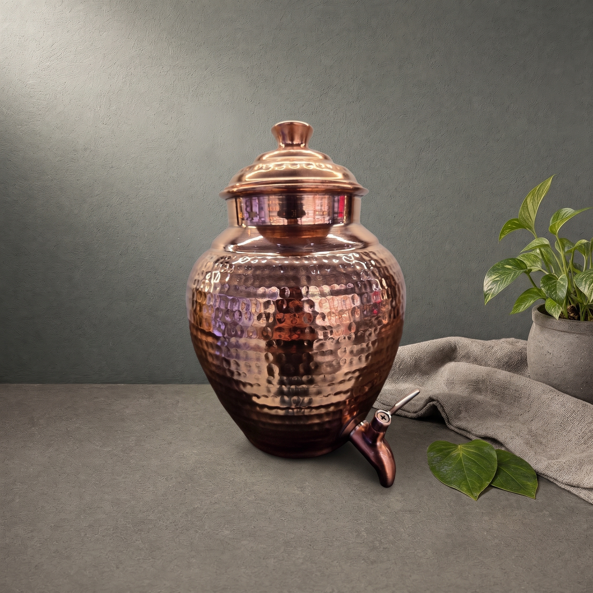 Hand Hammered Copper Water Dispenser on a stone ledge against a grey plaster wall - Peetal Parampara