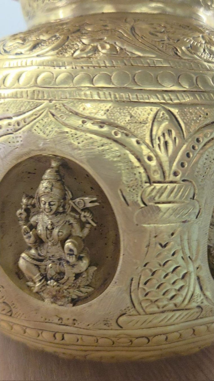 Close view of an engraved brass kalash, with a seated Lakshmi figure set into a recessed medallion on its body.