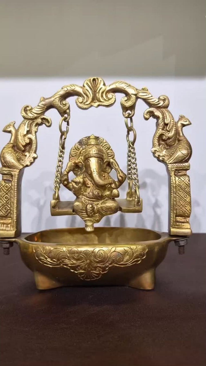 A brass oil lamp on a footed bowl, with a seated Ganesha under an arch flanked by facing peacocks.